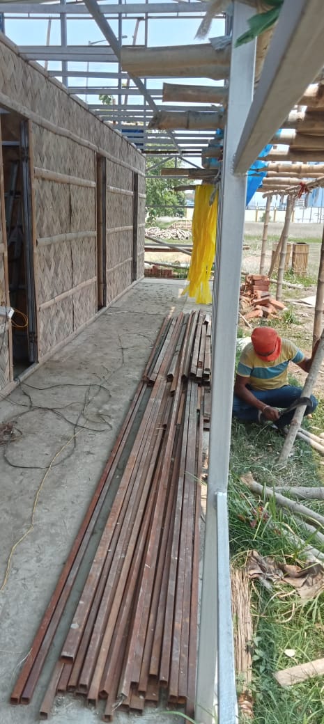 A karigar working on the veranda, iron beams stacked alongside, woven bamboo walls going up behind