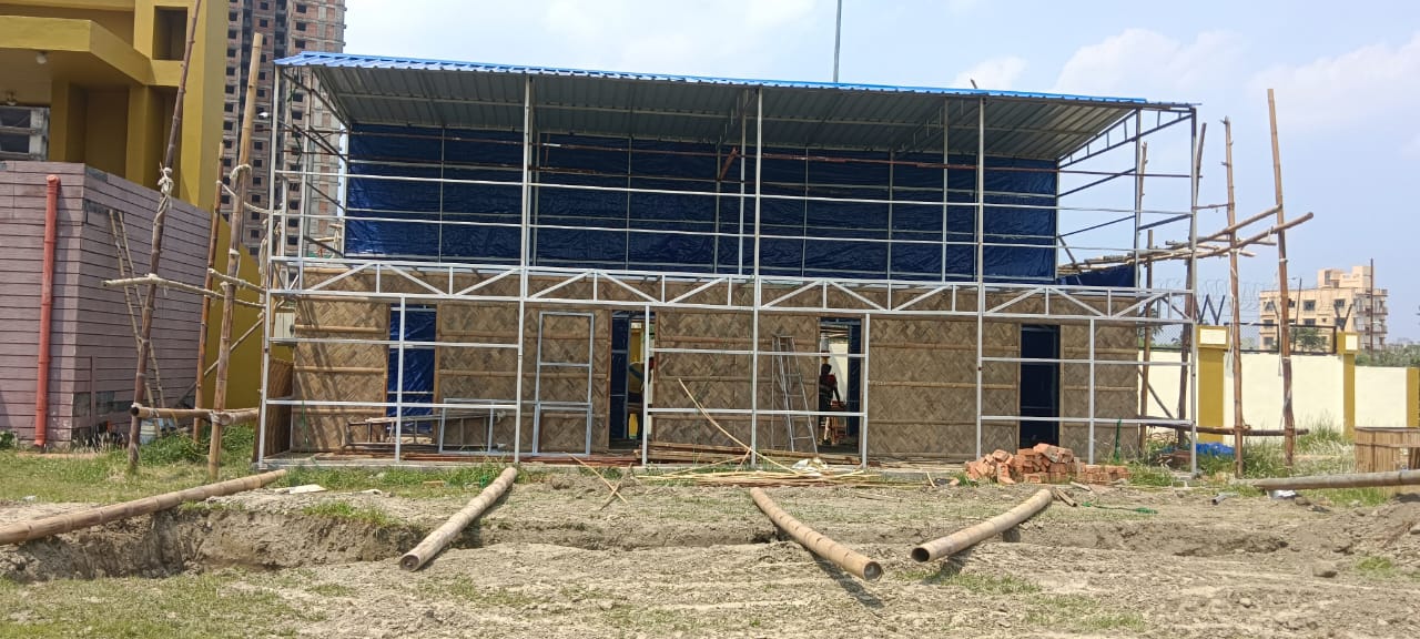 The bare steel framework with blue tarp roof, bamboo poles on the ground, woven panels beginning on the ground floor